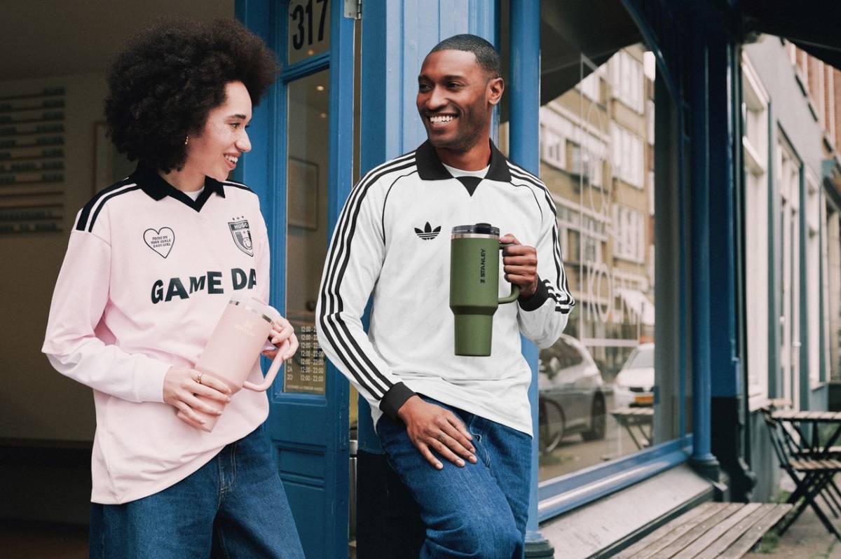 Жена с розова спортна блуза и мъж с бяла блуза adidas стоят пред синя врата на магазин и се усмихват, държайки термочаши Stanley Quencher H2.0 с дръжки в светлорозов и маслиненозелен цвят.