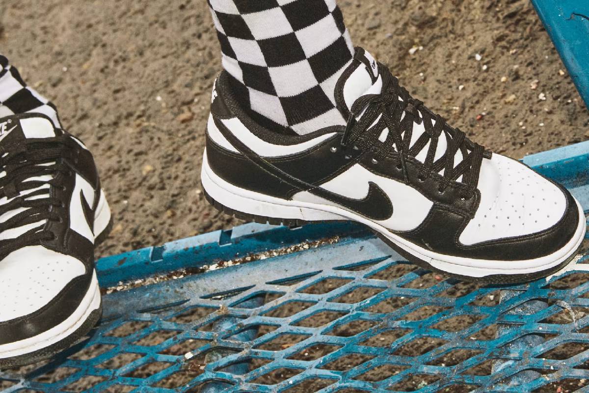 Близък план на спортни обувки Nike Dunk Low Retro в черно и бяло, комбинирани с високи карирани чорапи върху синя метална скара.