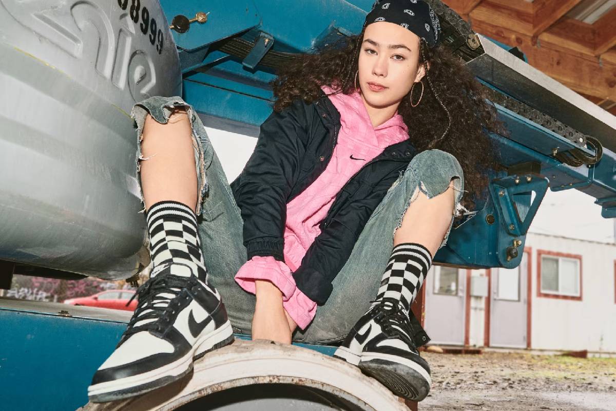 Жена с къдрава коса и бандана, облечена с розово худи Nike, черно яке и дънки, позира между части от камион. Акцент върху черно-бели маратонки Nike Dunk Low и карирани чорапи.“